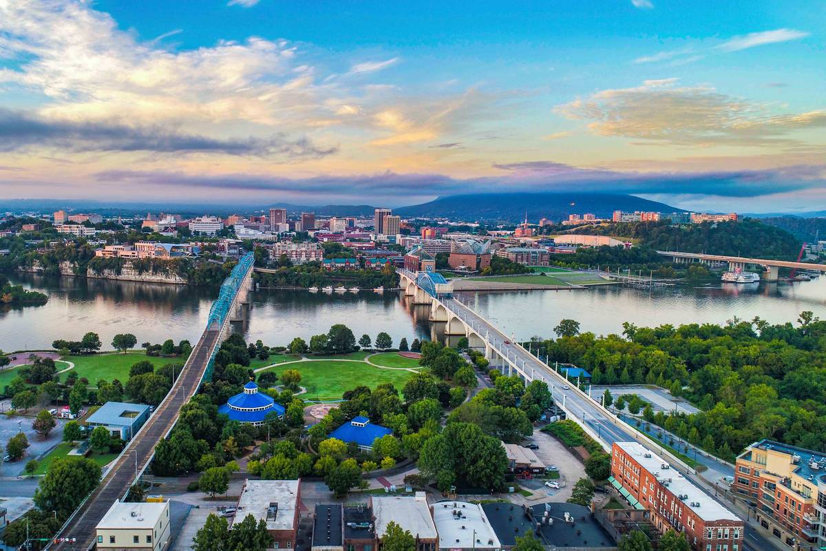 Elevated view over the Tennessee River in Chattanooga at dusk, with bridges crossing to downtown, riverfront parkland in the foreground, and a mountain ridge on the horizon.